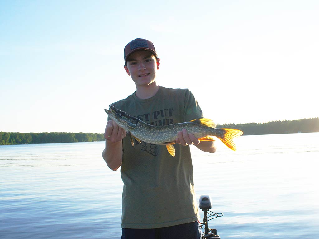 A teenaher in a cap holds a Pike on a boat by a calm lake, enjoying Minocqua fishing amid trees and a clear sky in the background.