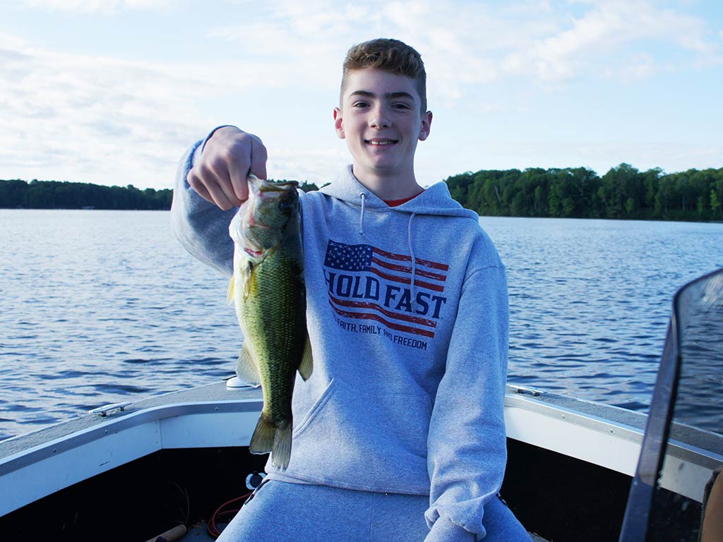 A teenager sitting in a boat on a lake near Minocqua holds up a freshly caught Smallmouth Bass and smiles toward the camera.