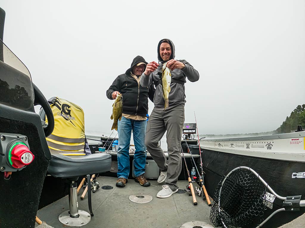 Two people in hoodies are standing on a fishing boat, proudly holding up the Bass and Walleye they caught during their Minocqua fishing trip. Their fishing rods and gear are scattered across the deck, showing they've had an active day out on the water.