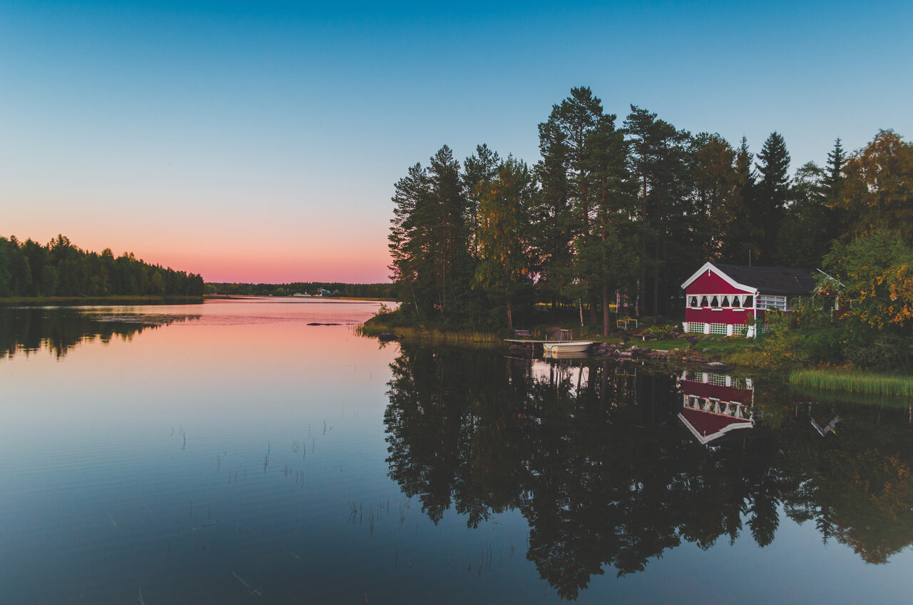 Blick &uuml;ber das Wasser bei Sonnenuntergang bei Umea. An Land rechts ist ein kleines rotes Haus zu erkennen, vor dem ein Boot im Wasser liegt.