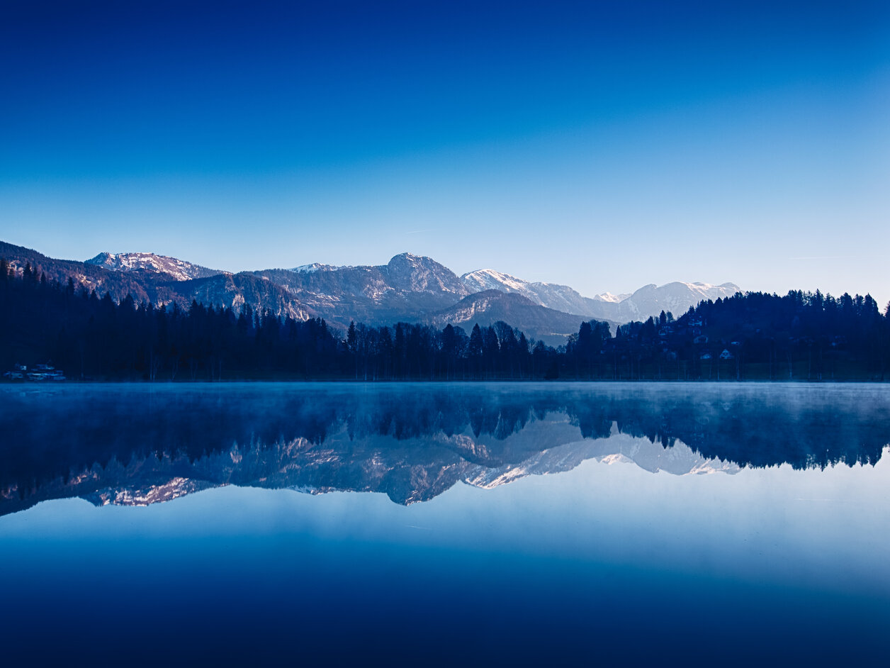 Die vordere H&auml;lfte des Bildes wird vom blauen Wasser des Putterersees eingenommen. Im Hintergrund sind die Alpen zu sehen.
