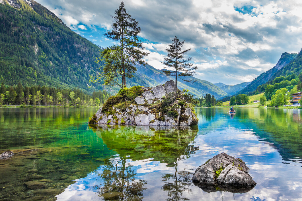 Blick auf einen gro&szlig;en Felsen im Hintersee, auf dem zwei Nadelb&auml;ume wachsen, mit den Alpen im Hintergrund.