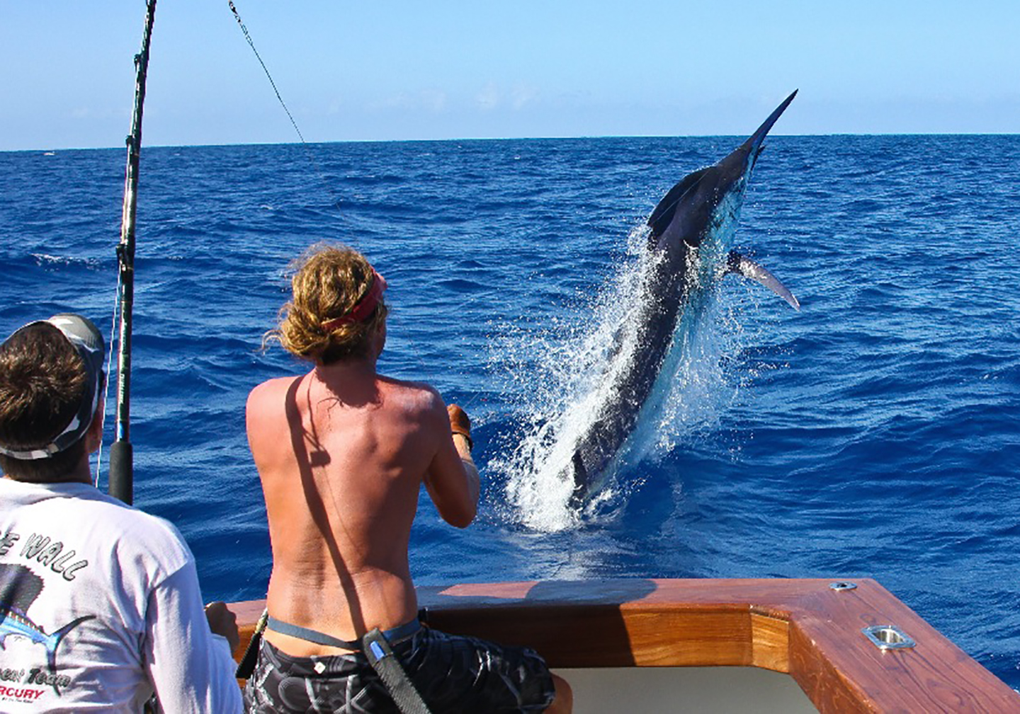 Ein m&auml;nnlicher Angler mit freiem Oberk&ouml;rper auf einem Angelboot, der mit einen gro&szlig;en Schwarzen Marlin k&auml;mpft, der aus dem Wasser springt.