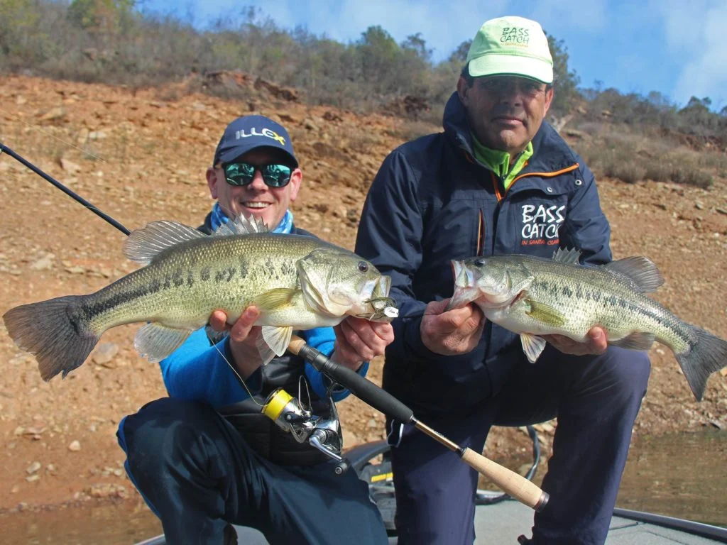 Zwei Angler halten zwei gro&szlig;e Forellenbarsche, mit dem Flussufer im Hintergrund.