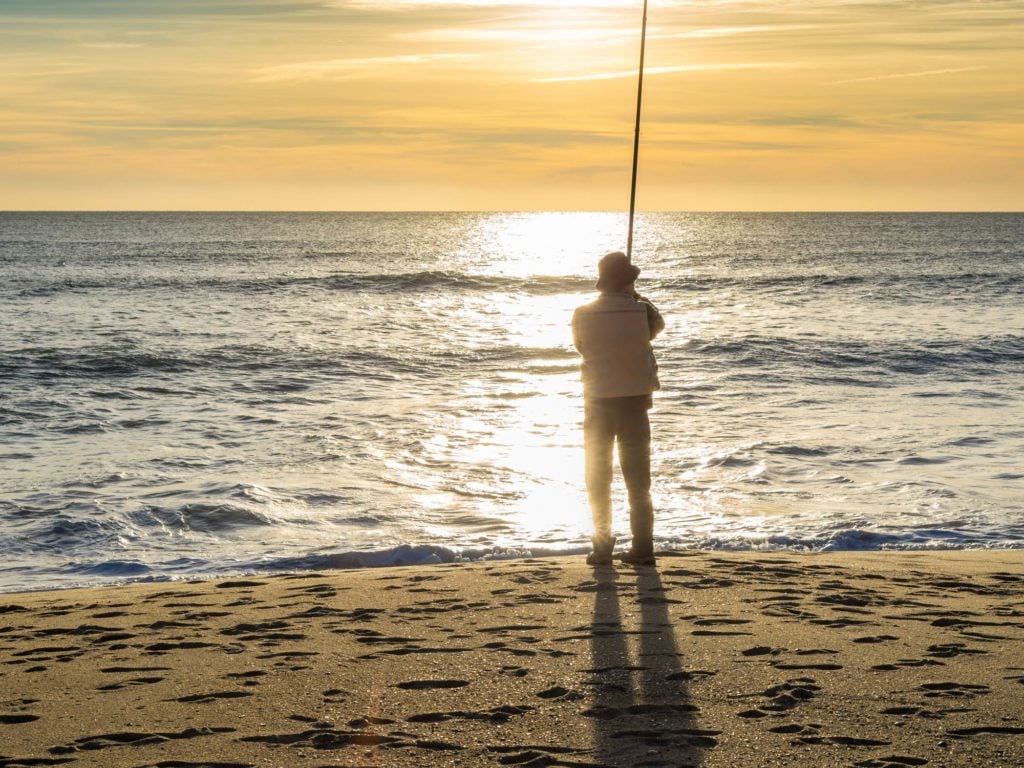 Ein einsamer Angler, der bei Sonnenuntergang auf einem Sandstrand steht und angelt.