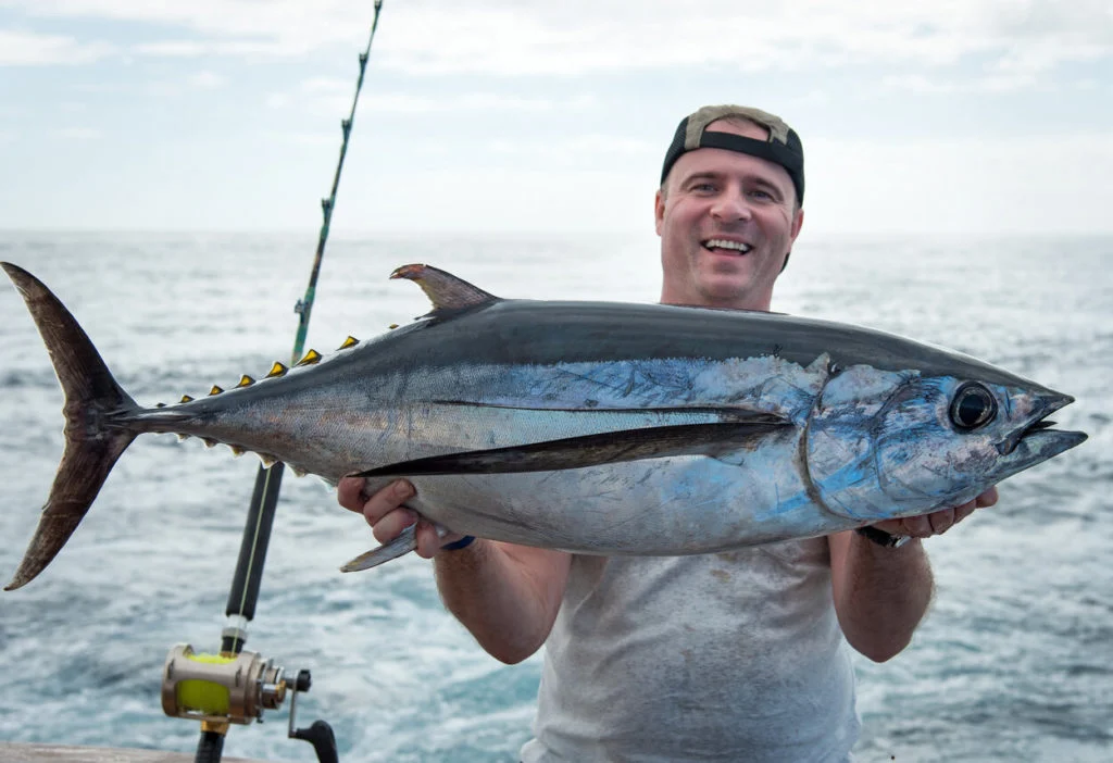 Ein fr&ouml;hlicher Angler, der auf einem Boot einen Blauflossen-Thunfisch in die Kamera h&auml;lt.