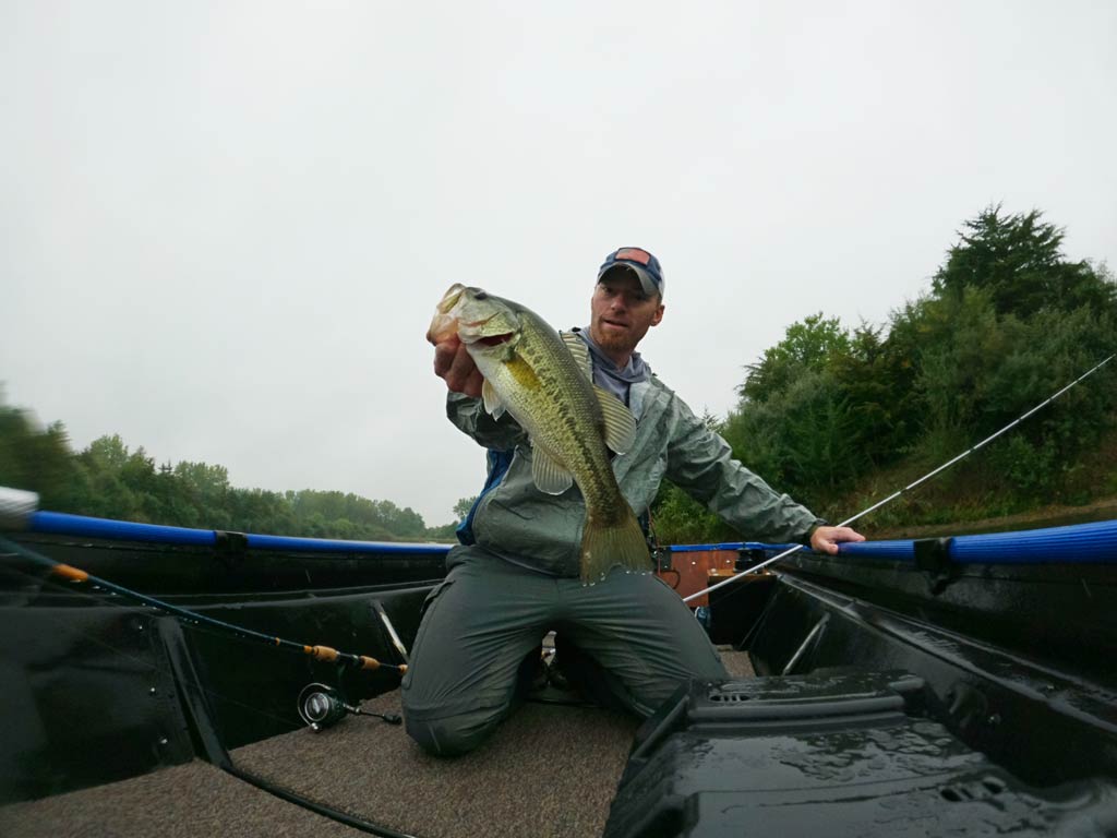 A man crouching on a small boat and holding up a Bass fish in one hand and a rod in the other on a cloudy day.