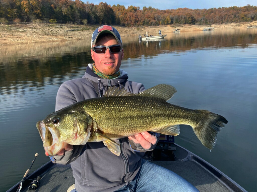 A man in a baseball cap holding a large Bass caught after using a front-facing sonar on a boat on a reservoir in Arkansas on a bright day.