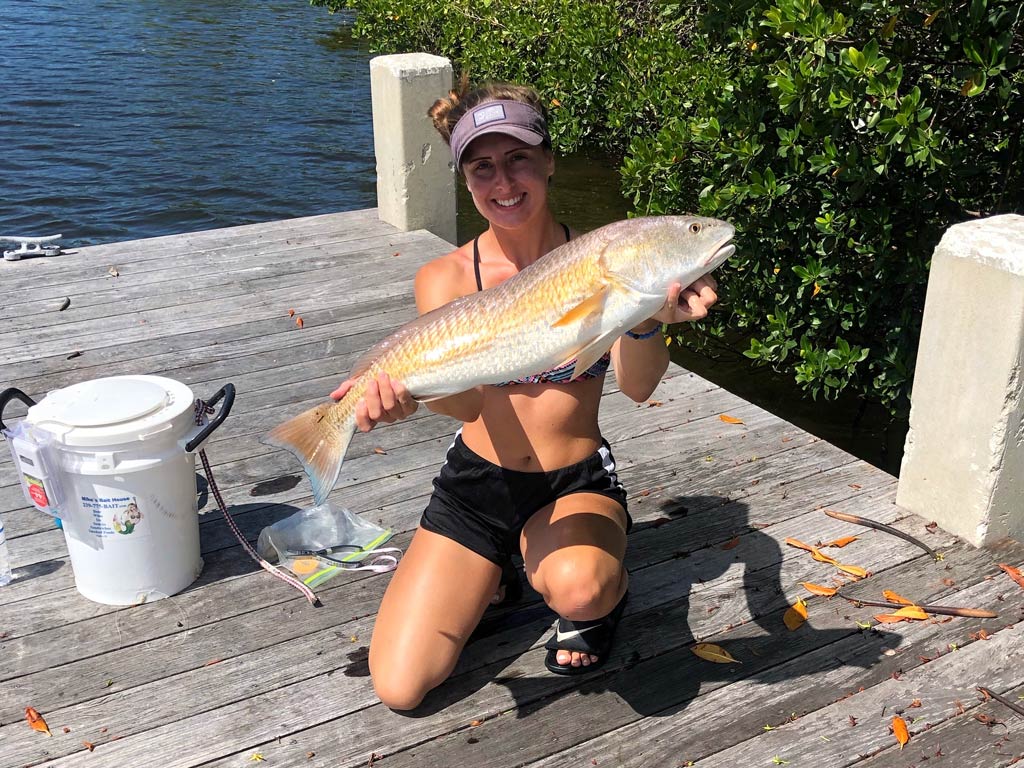 A woman in a cap, crouching on a dock next to a bucket and holding a large Redfish after a successful day solo fishing.