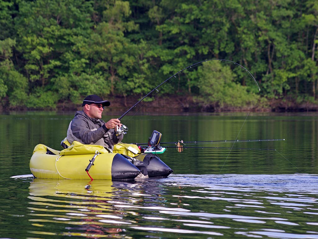 A man fishing from an inflatable yellow float tube on a calm lake, surrounded by trees, is using forward-facing sonar while he's holding a fishing rod with another rod beside him.