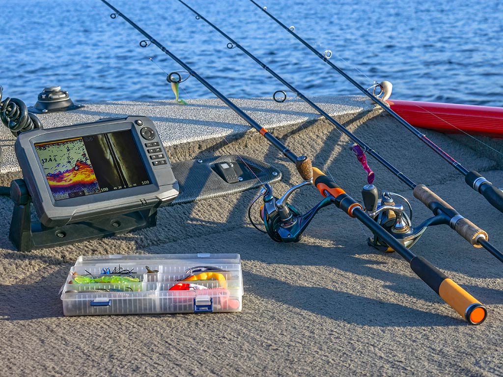 Three fishing rods, a tackle box with lures, a fishfinder with forward-facing sonar, and a smartphone are arranged on a boat's surface by the water.