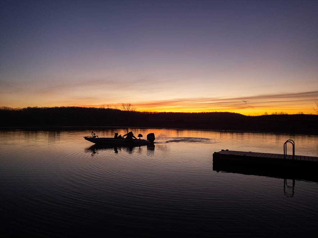 A view towards a small boat with two people as it glides across a calm lake at sunrise. They're leaving ripples near a dock in the foreground. You can see the silhouettes of the boaters as they search for Bass or Walleye hiding among submerged logs beneath the glassy surface. 