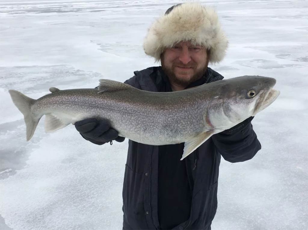 A person wearing a fur hat and gloves is holding a large Lake Trout on a frozen, snow-covered surface in Vermont.