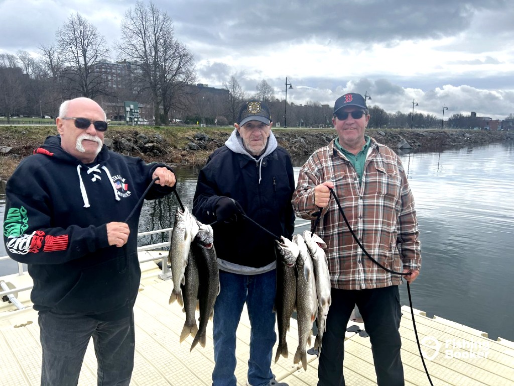 Three men stand on a dock by the water, each holding several large Salmon they've caught. Trees and buildings are visible in the background under a cloudy sky. T