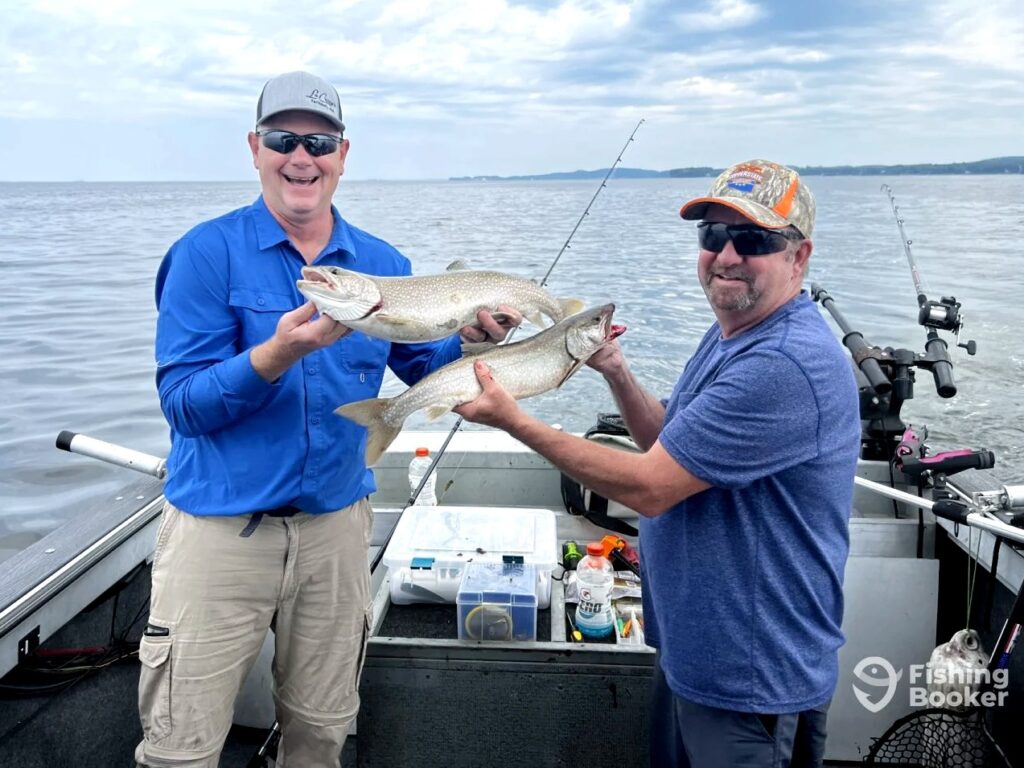 Two men are standing on a boat, each holding a large Lake Trout. Fishing gear is scattered around their feet, and they're surrounded by calm water stretching into the distance. The sky above is partly cloudy, adding depth to the scene.