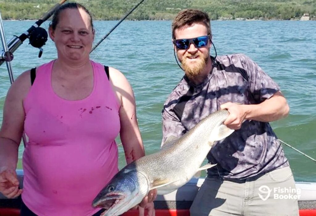 Two people are on a boat holding a large Lake Trout, with a lake and trees in the background. The person on the right is wearing sunglasses and holding the fish.