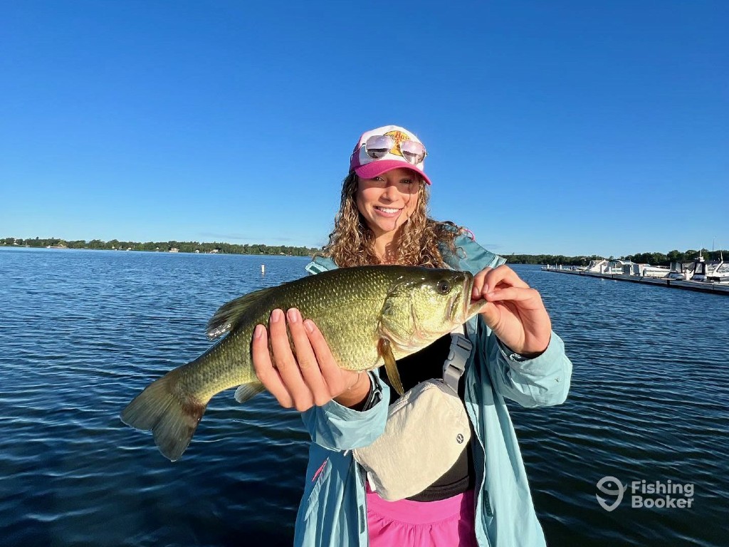 A young woman standing on a boat, holding a Smallmouth Bass with both hands and smiling. In the background, you can see a lake and a marina, reflecting the summer fishing season in Vermont.