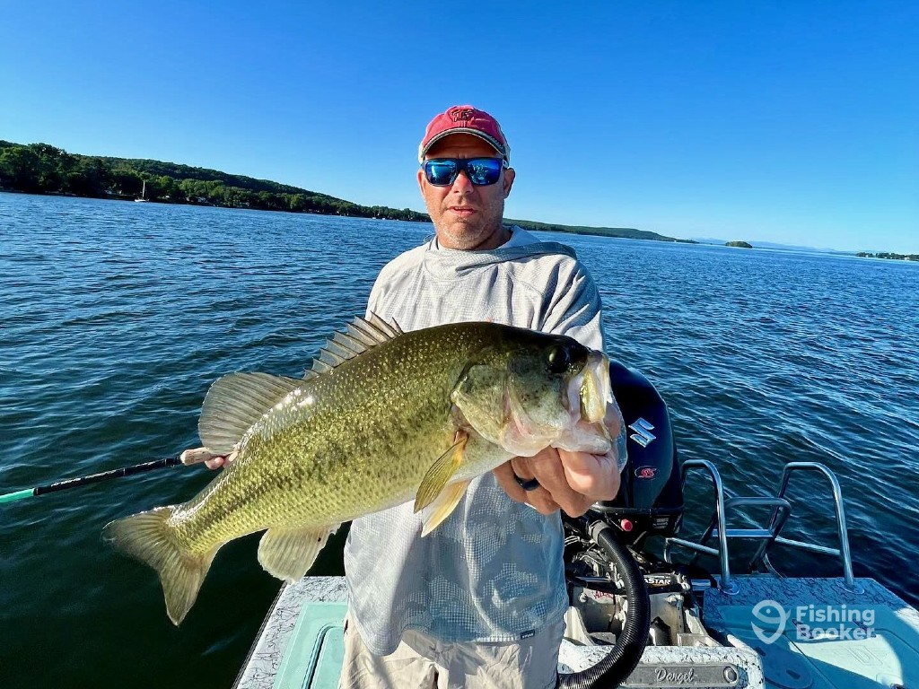 A man on a boat holds a large Bass with one hand, while standing on a lake under a clear blue sky, showing summer fishing in Vermont.
