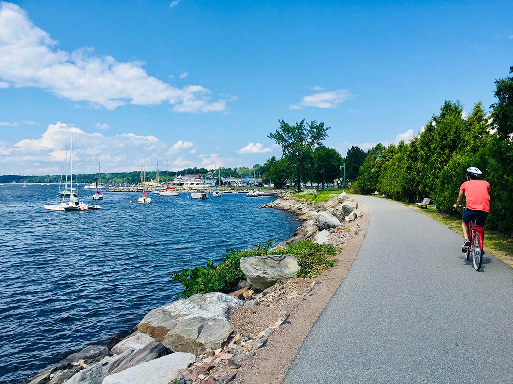 A view from behind of a person riding a bicycle on a paved path beside a body of water in Vermont with sailboats visible, under a blue sky with scattered clouds.