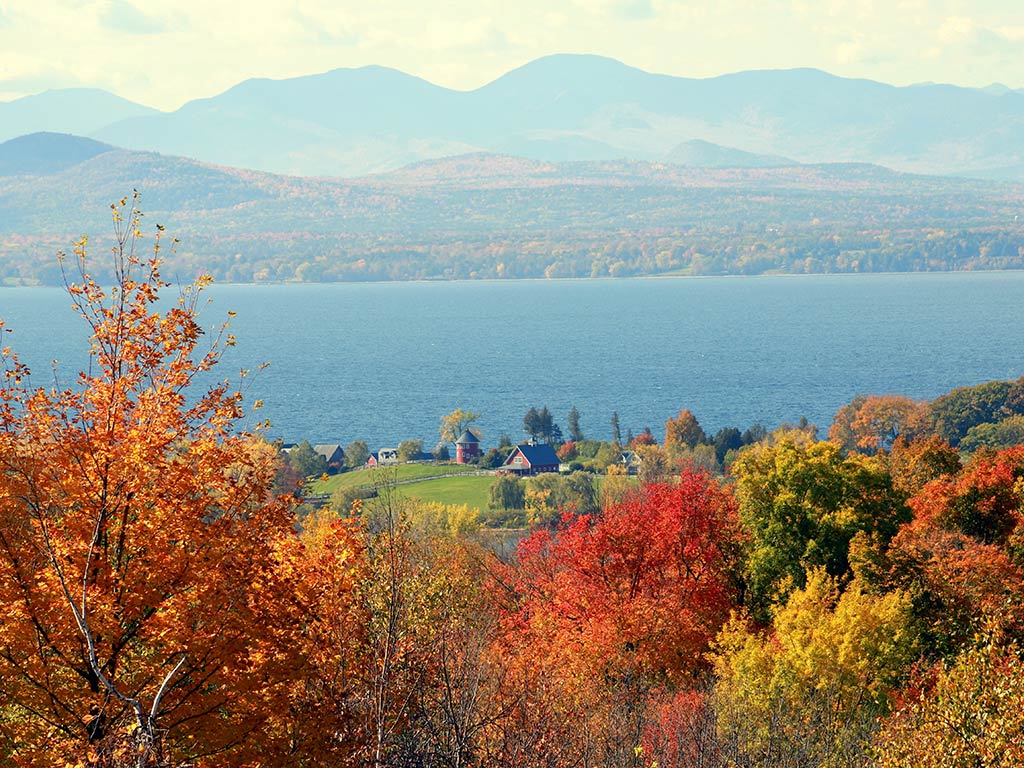 Colorful autumn trees frame a lake with distant mountains and a small cluster of buildings visible near the shoreline under a hazy sky in Vermont.