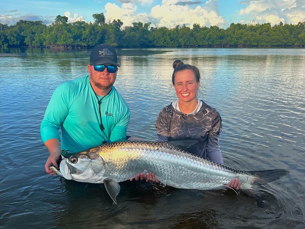 Two anglers are standing in shallow inshore water, holding a large Tarpon. There are trees and a cloudy sky in the background.