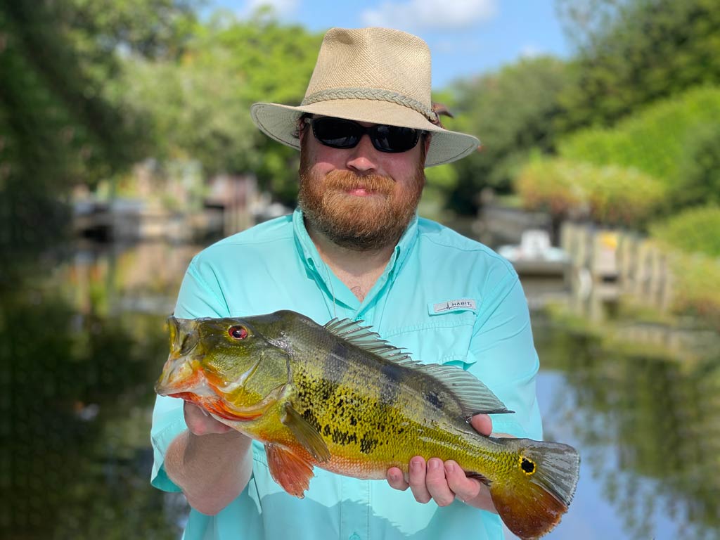 Drew Pawley, FishingBooker’s most passionate angler, wearing a hat and sunglasses while posing with Peacock Bass in Florida near the dock.