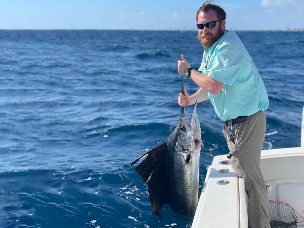 Drew Pawley, FishingBooker’s most passionate angler, posing on a deep sea fishing charter while holding a large Sailfish vertically over the side of the boat.