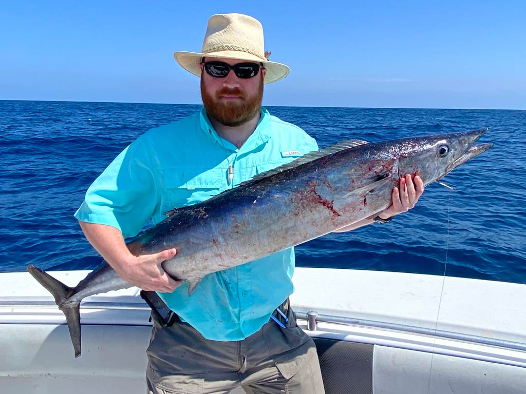 Drew Pawley, FishingBooker’s most passionate angler, standing on a charter boat and wearing a hat and sunglasses posing with a Wahoo he caught while fishing offshore.