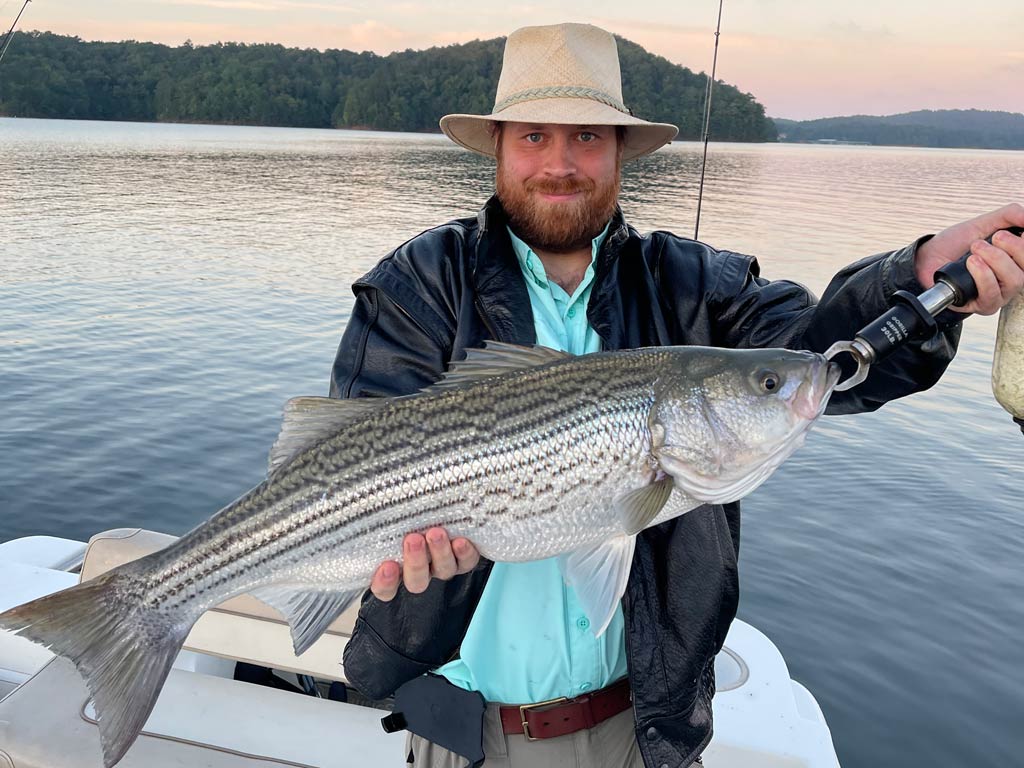 Drew pawley wearing a hat and leather jacket, holding a large Striped Bass on a boat with a calm lake and forested shoreline in the background.