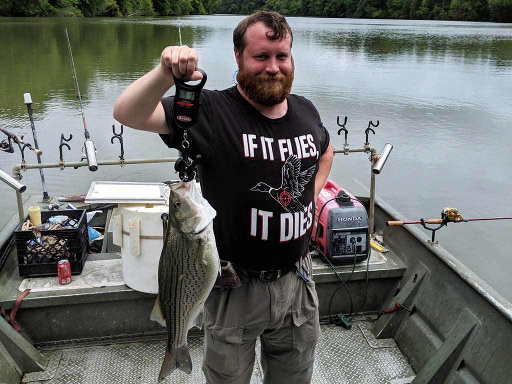 Drew Pawley on a fishing boat, holding up a large Bass with a scale. Fishing rods and equipment are around him, with a lake and trees in the background.