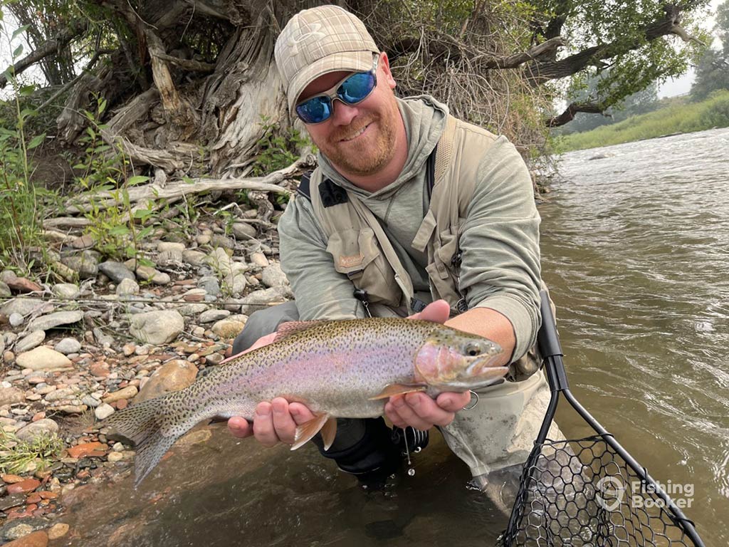 A man kneels by a riverbank holding a large Trout, wearing fishing gear, sunglasses, and a cap. There's a fishing net on the ground beside him.