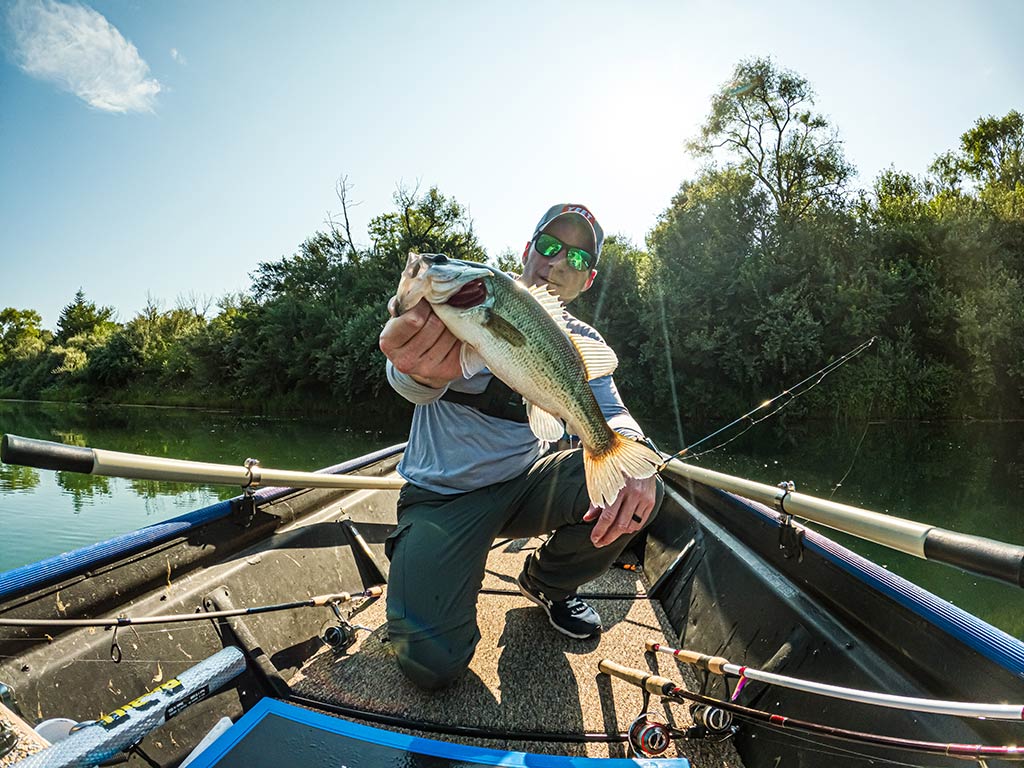 A man on a boat holding a Bass toward the camera, with fishing rods and gear in view against a wooded shoreline in the background, somewhere in Wisconsin.