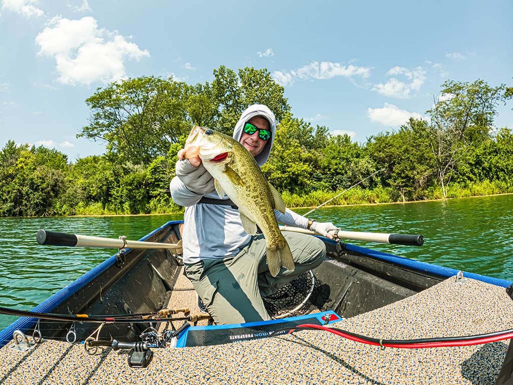 Someone's sitting in a boat on a lake, proudly holding up a big Wisconsin Bass with one hand and a fishing rod in the other. They're surrounded by trees and a blue sky.
