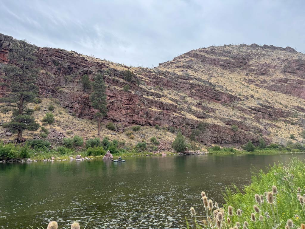 A calm river flows beside a rocky, shrub-covered hillside under a cloudy sky, with someone in a boat near the riverbank.