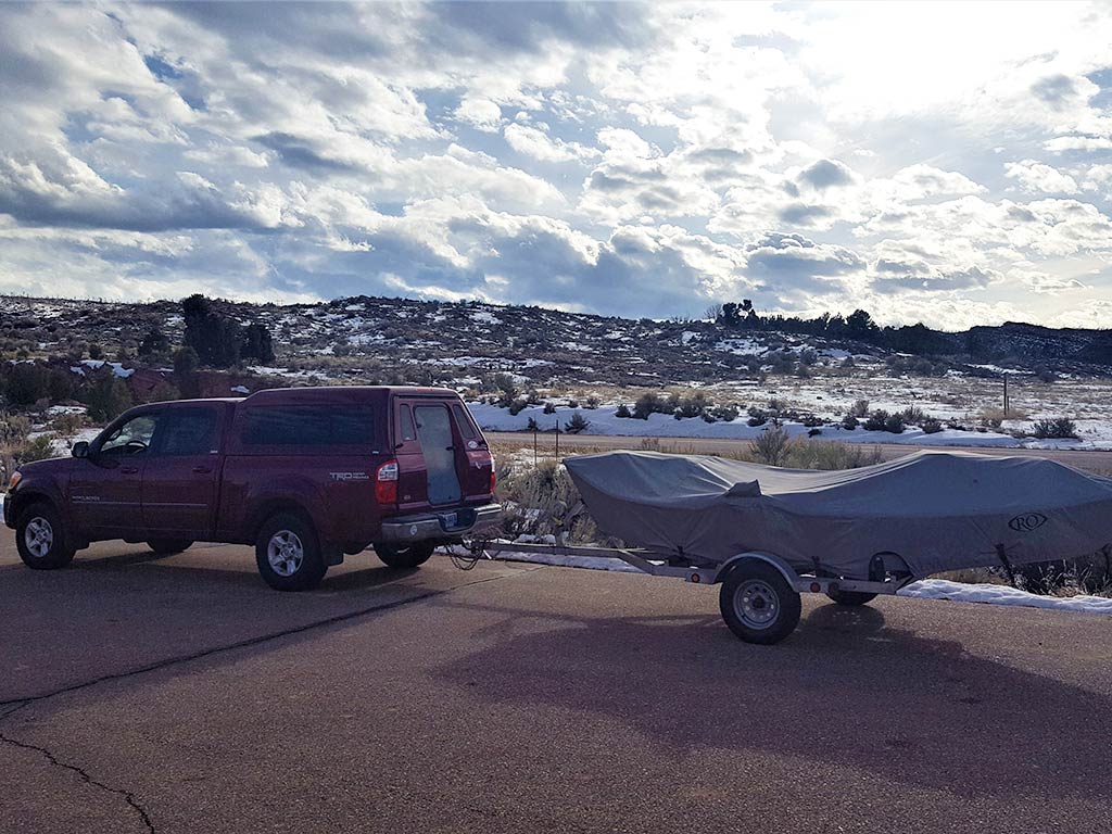 A maroon Toyota Tundra on a paved road, towing a covered boat trailer in front of snow-dusted hills near the Green River.