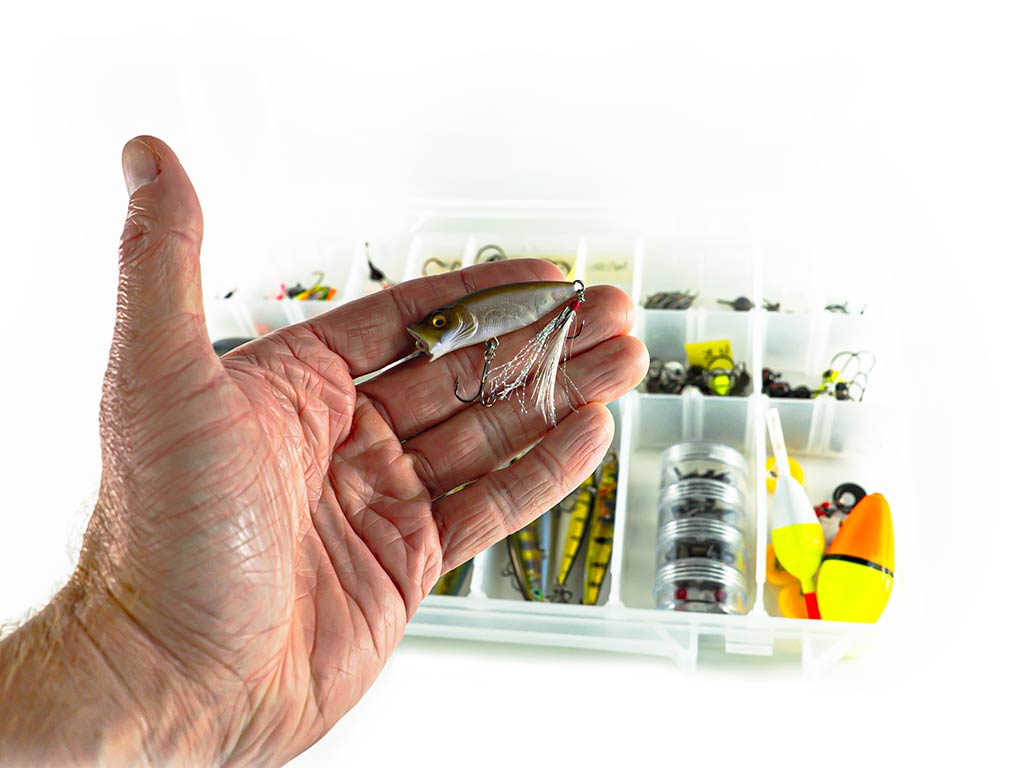 A hand’s holding a small fishing lure, with a tackle box of fishing gear—perfect for targeting Wisconsin Bass—in the background.