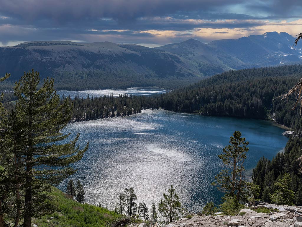 A scenic view of Lake Mary in Mammoth Lakes shows a sparkling lake surrounded by tall Pine trees and rugged Sierra Nevada Mountains beneath a cloudy sky.