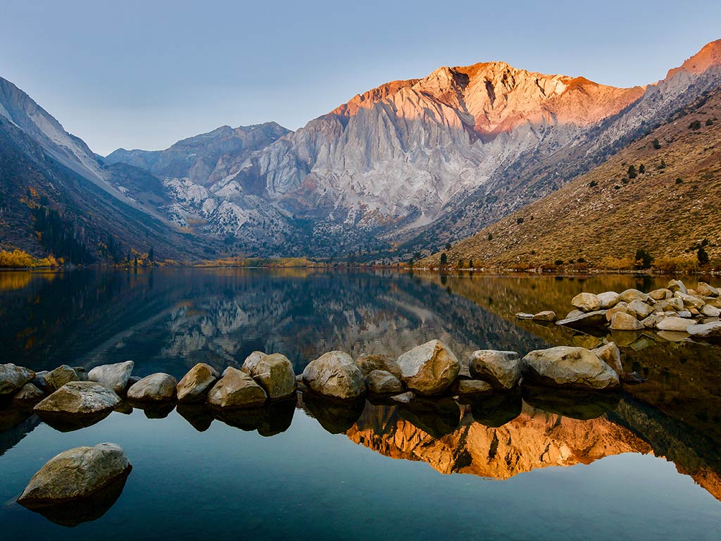 A calm lake reflects rocky mountains at sunrise, with a line of stones in the foreground under a clear sky.