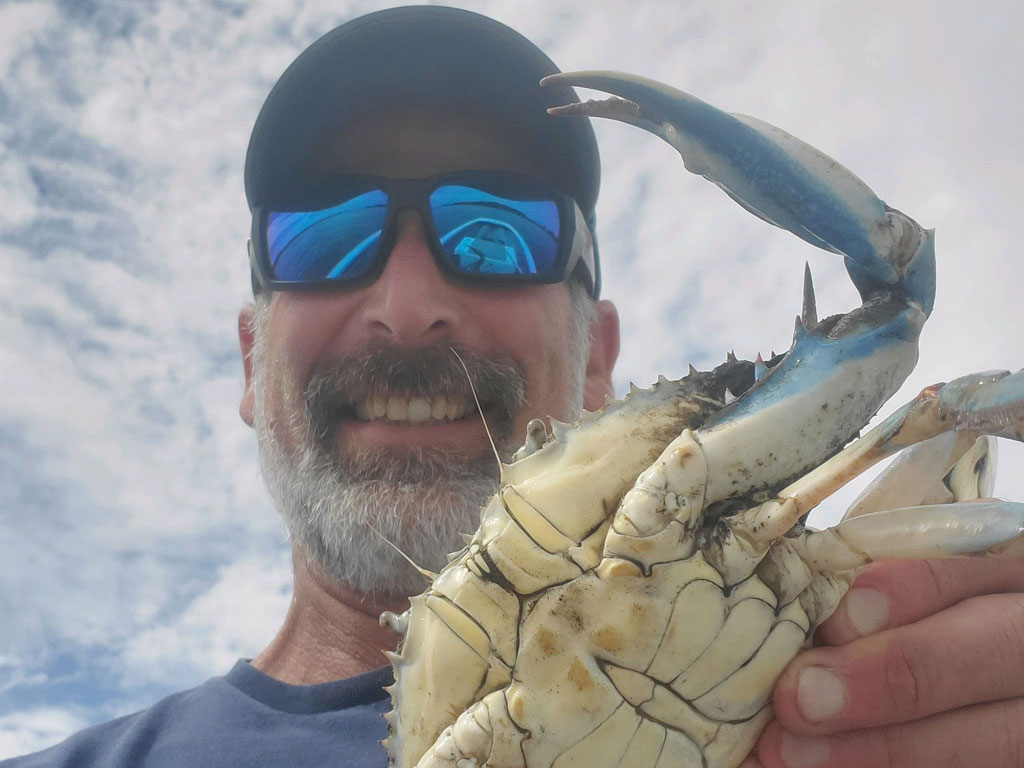 Amiling man wearing sunglasses and a cap holds up a large Blue Crab with one claw raised, set against a cloudy sky background.