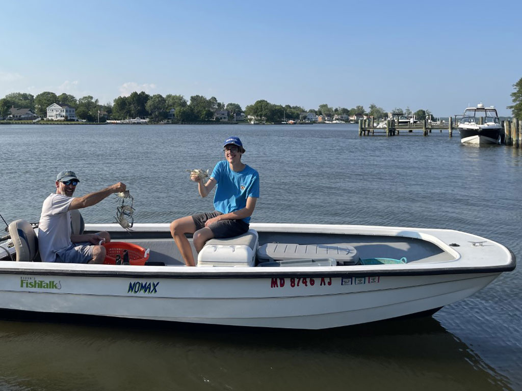 Two people sit in a small motorboat on the calm waters of the Chesapeake Bay. One is holding up a Crab, while and houses and docks are visible behind them under a clear sky.