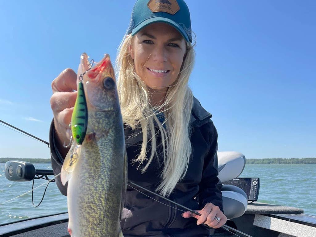 A woman holding a freshly-caught summer Walleye with a lure attached, sitting on a boat on a lake under a clear blue sky.