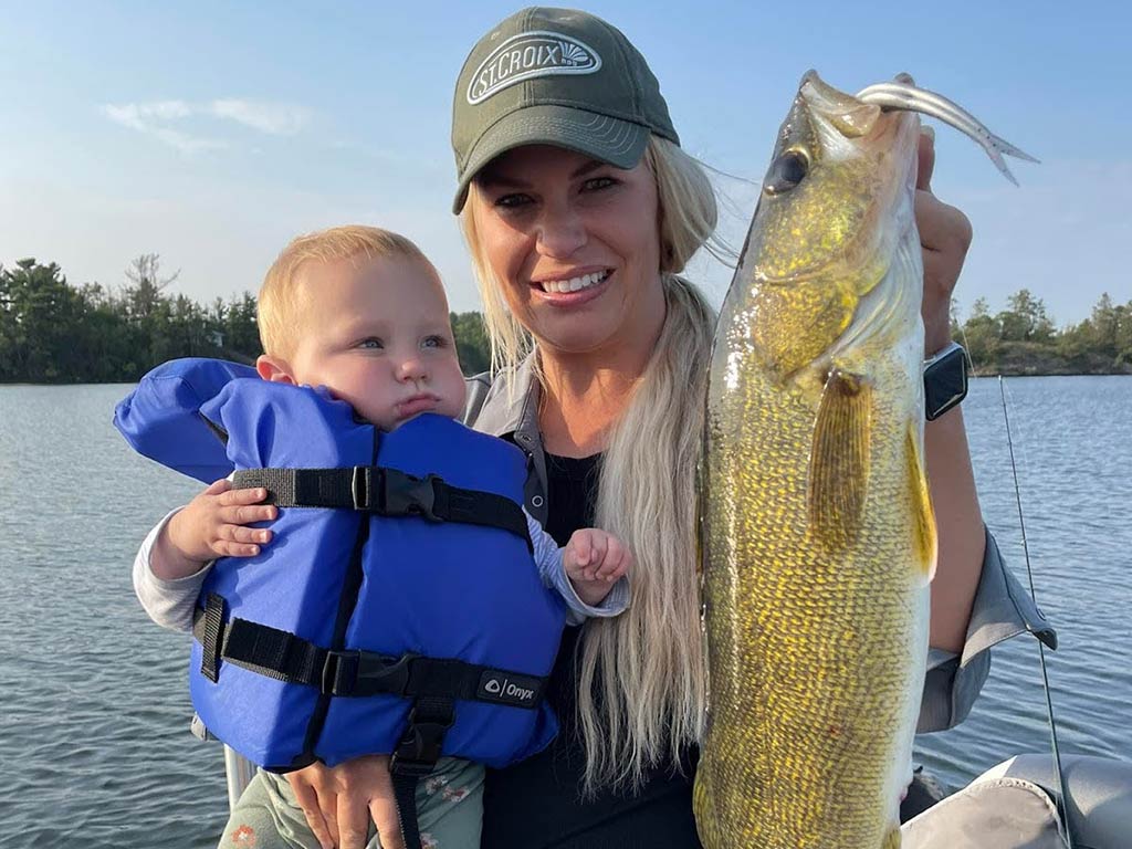 A woman in a cap holding a large summer Walleye in one hand and a baby in a blue life jacket in the other while on a boat on the water.