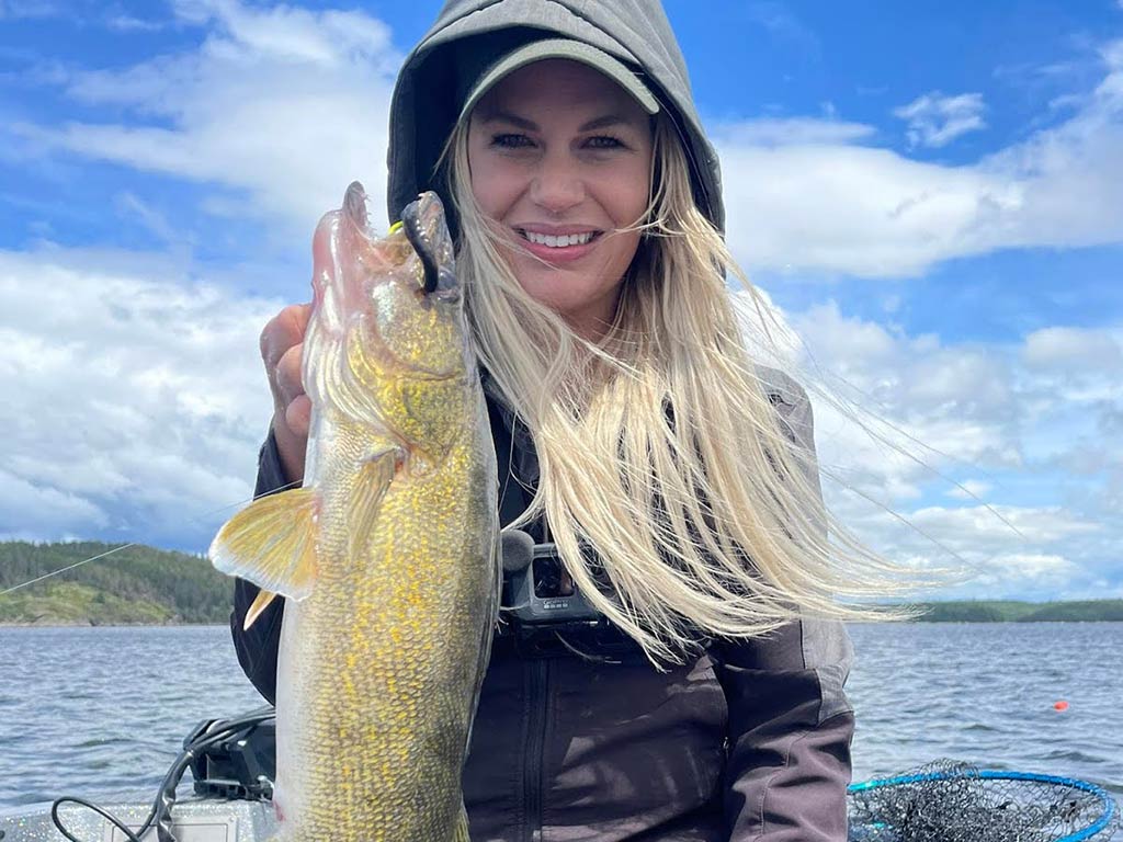 A woman in a hooded jacket holding a large summer Walleye on a boat, with a lake and cloudy sky in the background.