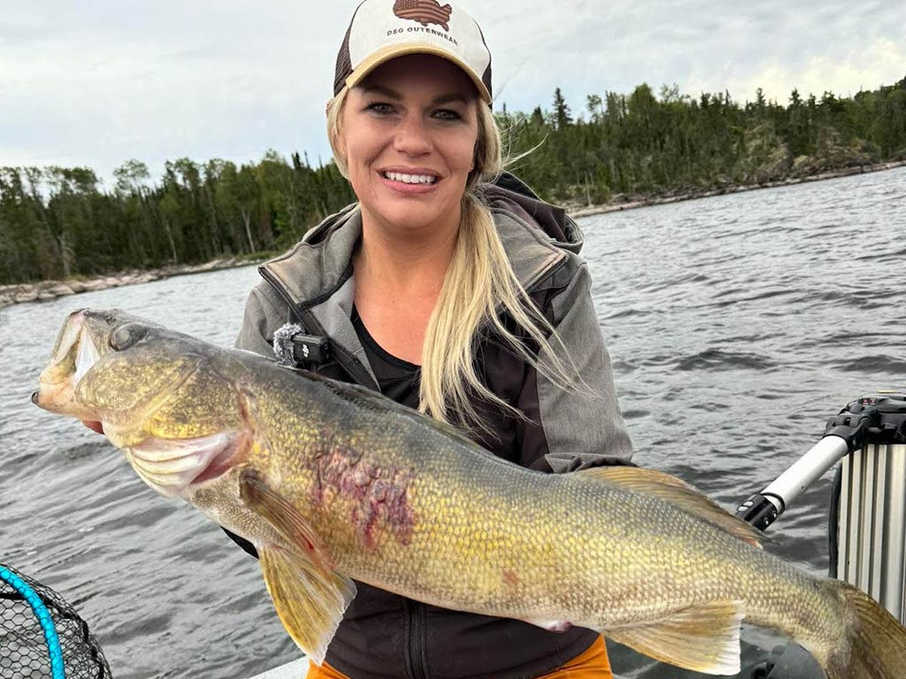 A woman on a boat holding a large summer Walleye with visible marks on its side; trees and water are in the background under a cloudy sky.