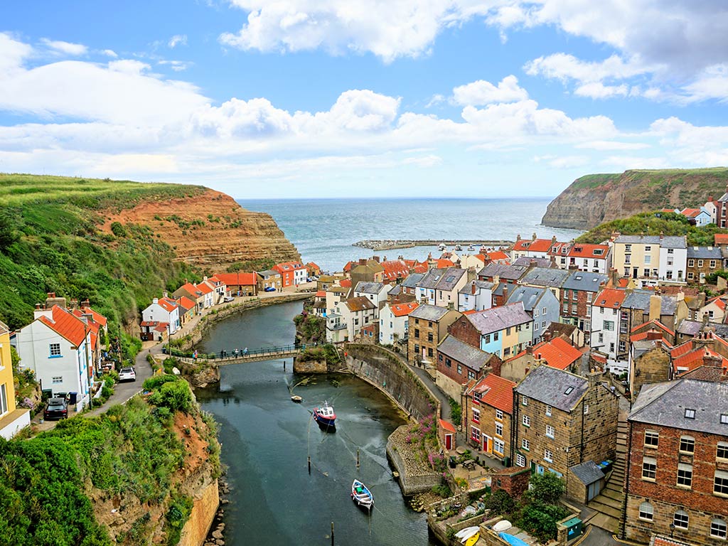 An aerial view of a coastal village in the UK with colourful houses, a winding river, small boats, a stone bridge, and cliffs by the sea under a partly cloudy sky.
