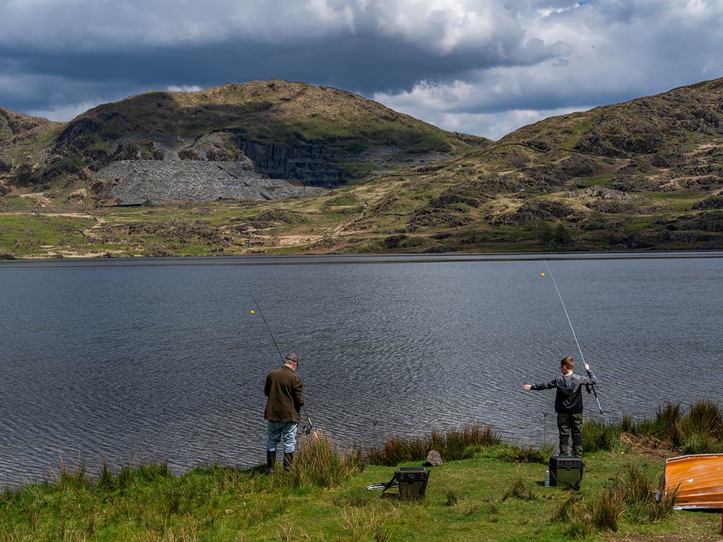 Two people are fishing with rods by the edge of a lake, with grassy hills and rocky slopes in the background under a partly cloudy sky. No fish like Bass or Trout are visible.