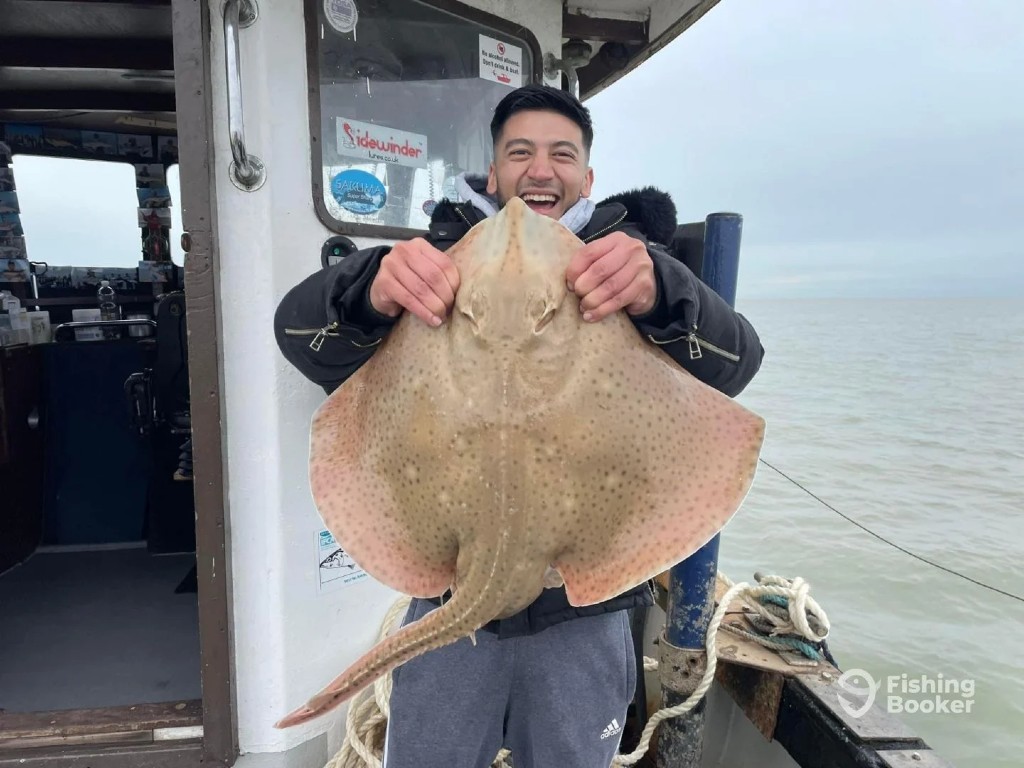 A man standing on a boat, holding up a large Skate fish and smiling at the camera. The sea is visible in the background on a cloudy day, typical of the UK no matter the season.