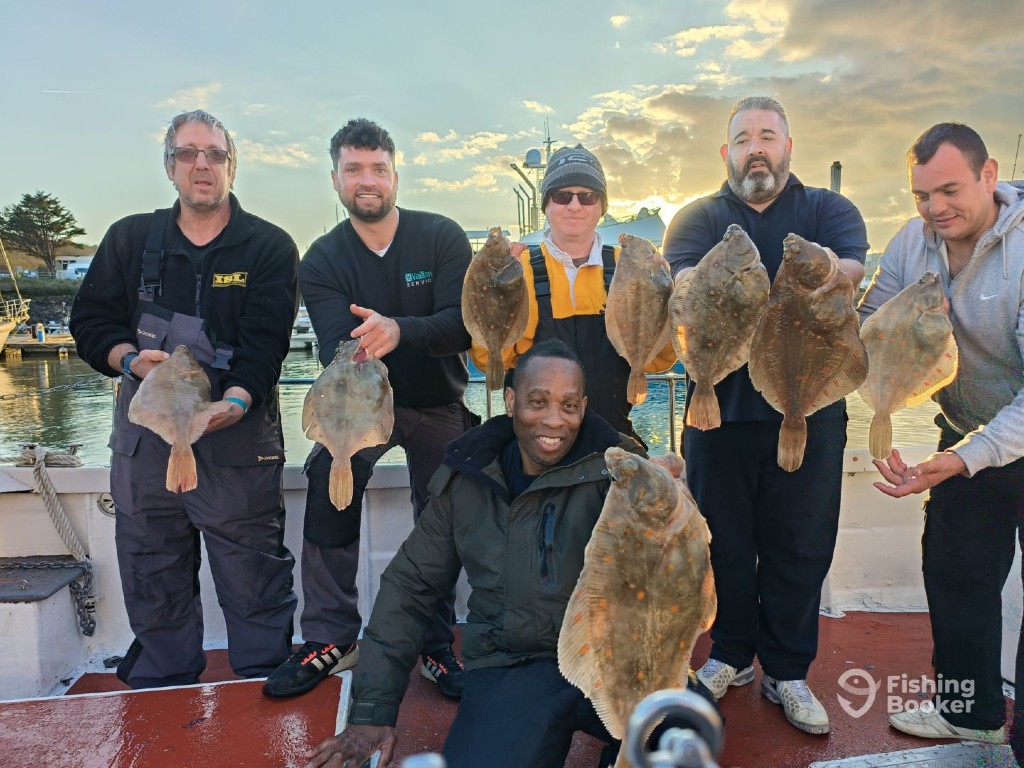 Five people are standing and one person is kneeling on a boat in the UK, each holding a large Flatfish, with the sea and cloudy sky in the background.