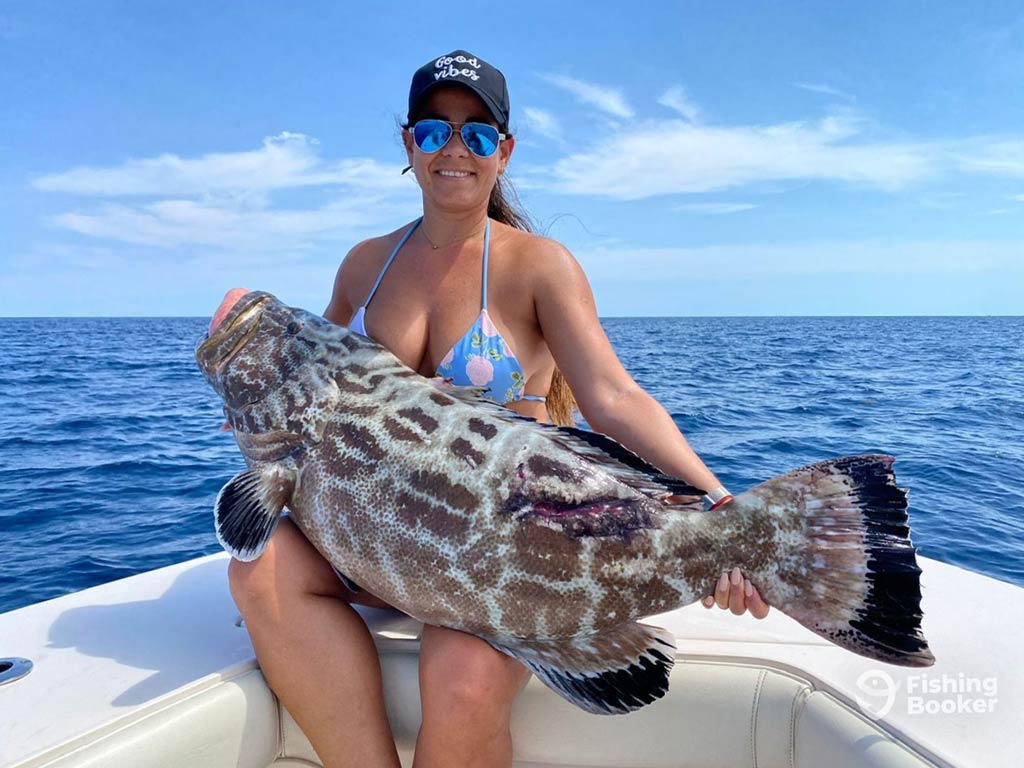 Woman's sitting on a boat holding a large Black Grouper, species of shallow-water complex that will be included in the Grouper Rule Changes soon, with the ocean and blue sky in the background.