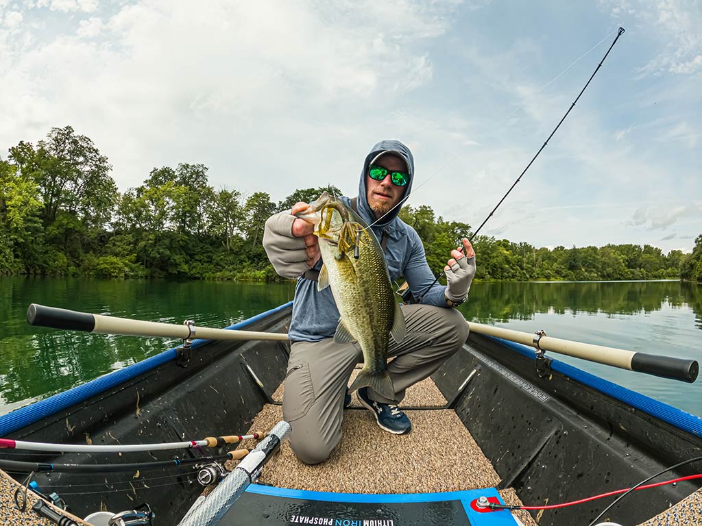 A person in fishing gear kneeling in a boat, holding up a Bass with one hand and a fishing rod with the other, surrounded by water and trees, showing off the that he caught using the free rig.